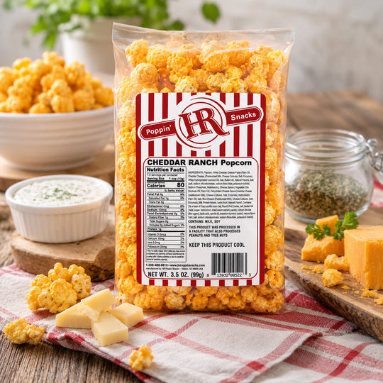 A clear bag of Poppin' Ranch Cheddar Ranch Popcorn sits on a red-and-white checkered cloth on a wooden table, surrounded by cheese wedges, a small bowl of dipping sauce, dried herbs, and extra popcorn. The product packaging is visible and the brand name Poppin' Ranch is readable on the front label.