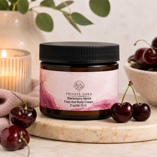 A jar of Private Oaks Blackberry Merlot Face and Body Cream sits on a marble slab surrounded by fresh cherries, with a softly blurred background featuring a candle and green leaves.