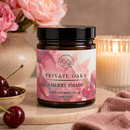 A jar of Private Oaks Cherry Smash Face and Body Cream sits on a marble board in a warm, styled setting with pink flowers, fresh cherries, and a lit candle in the background.