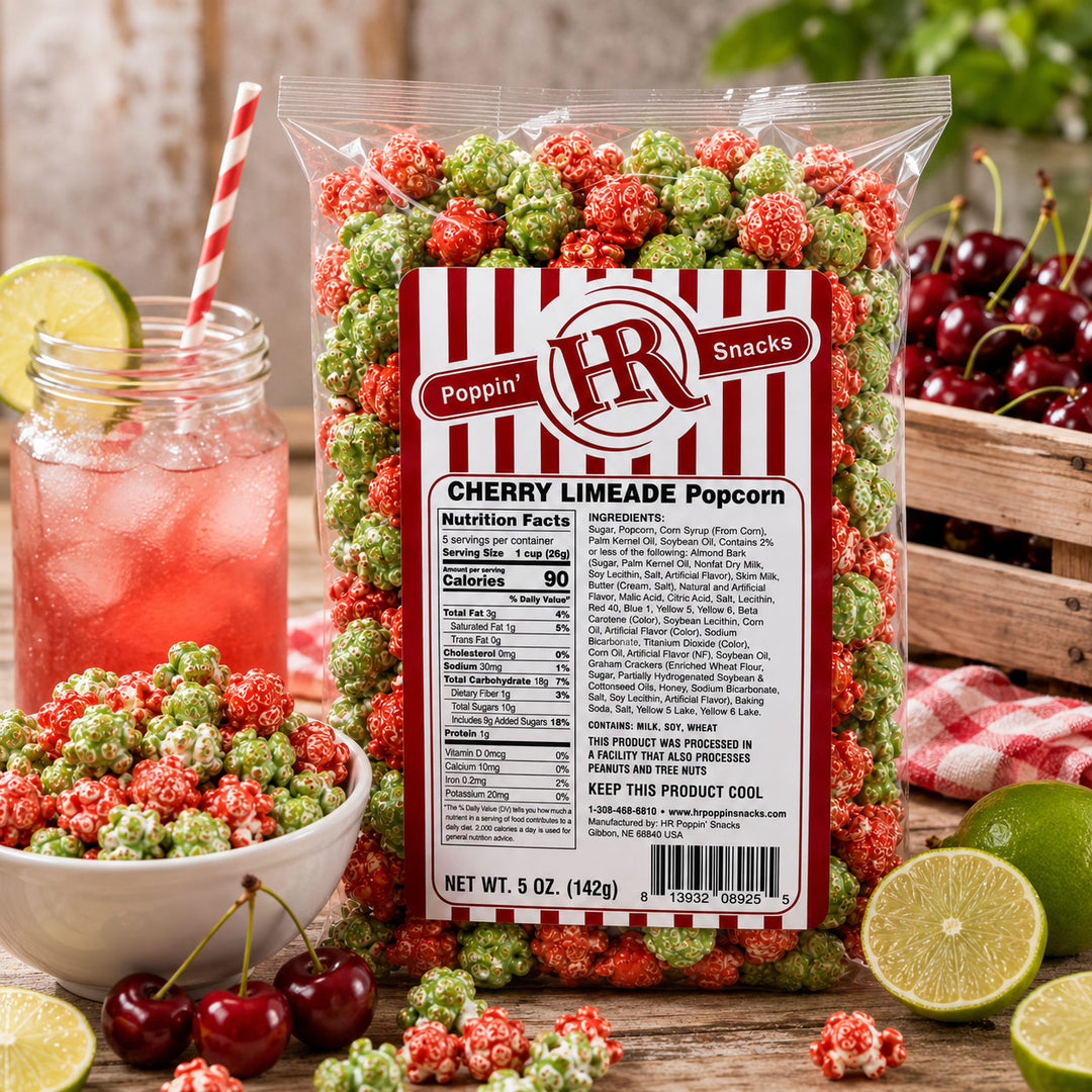 A 5 oz bag of Poppin' Snacks Cherry Limeade Popcorn in a clear bag with a red and white striped label, surrounded by cherry limeade, limes, cherries, and popcorn on a rustic wooden table.