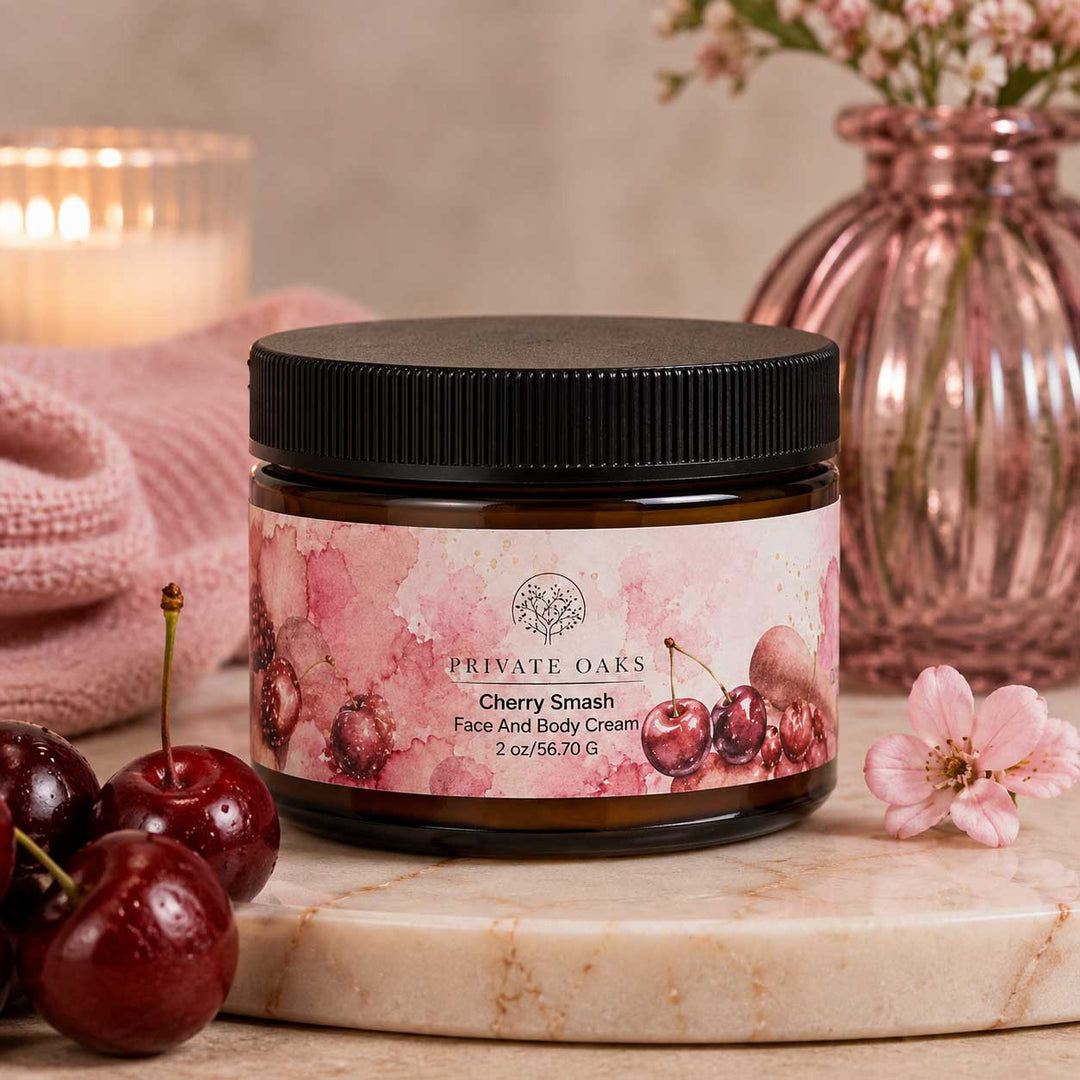 A jar of Private Oaks Cherry Smash Face And Body Cream sits on a marble slab, surrounded by fresh cherries, a pink knit blanket, and a pink glass vase in the background.