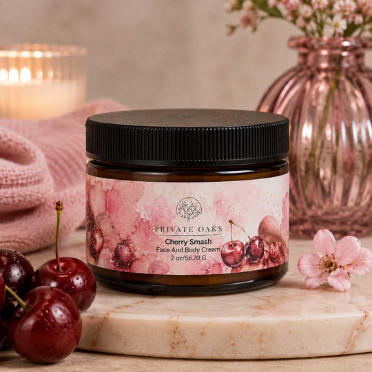 A jar of Private Oaks Cherry Smash Face And Body Cream sits on a marble slab, surrounded by fresh cherries, a pink knit blanket, and a pink glass vase in the background.