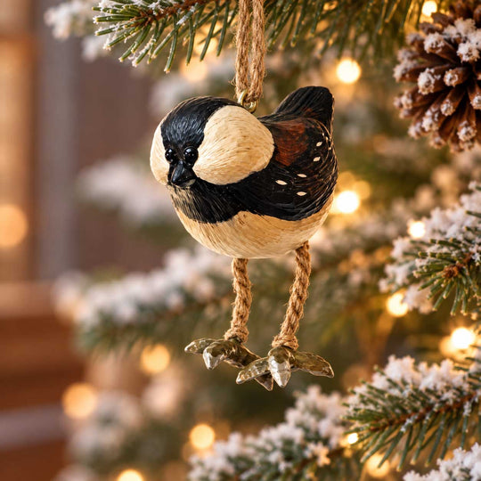 A bird-shaped Christmas ornament hanging from a pine branch on a decorated tree. The ornament is painted like a chickadee with a cream face and chest, black head, and brown wing patches, with rope legs and a metallic silver clawed foot hook.