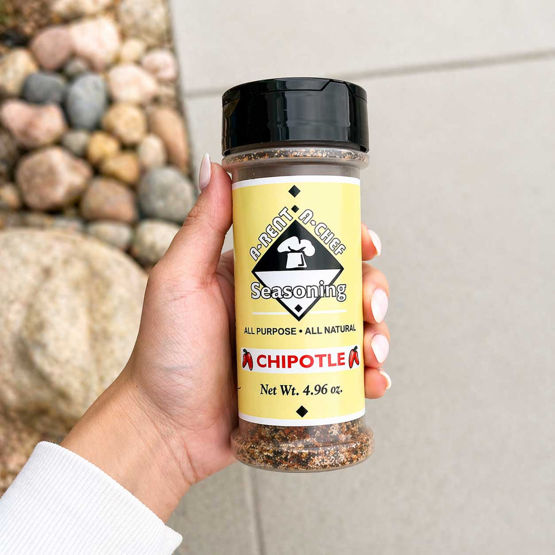 Hand holding a container of 'A-Rent-A-Chef Chipotle' seasoning with a natural stone background