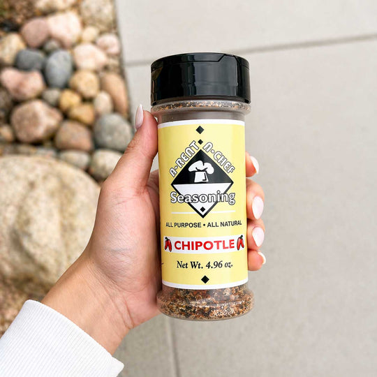 Hand holding a container of 'A-Rent-A-Chef Chipotle' seasoning with a natural stone background