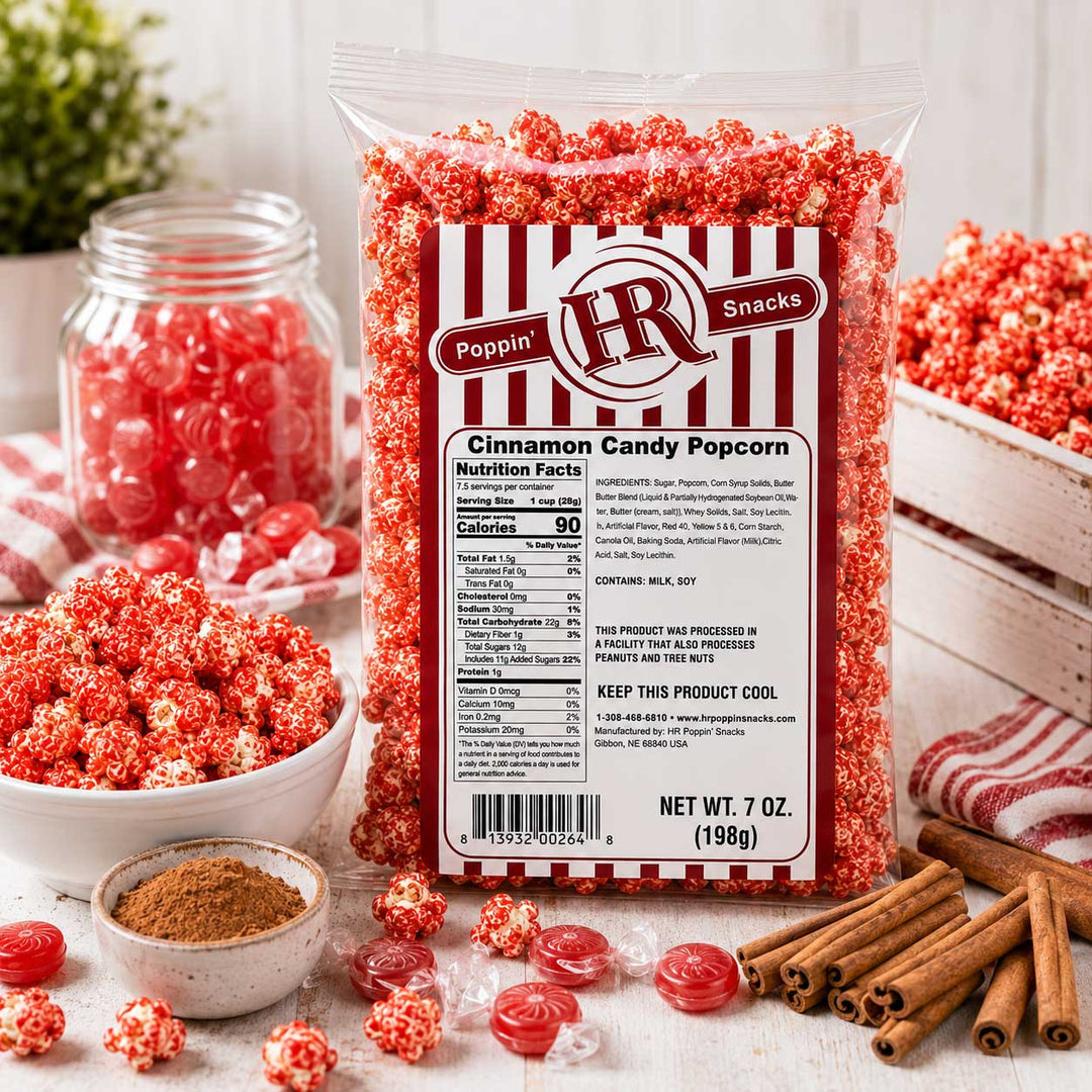 A 7 oz bag of Poppin' Snacks Cinnamon Candy Popcorn stands in a styled tabletop setup with bowls, a jar, cinnamon sticks, and loose candy popcorn pieces. The bag has a red-and-white striped label with product details and a QR code.