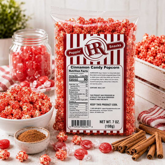 A 7 oz bag of Poppin' Snacks Cinnamon Candy Popcorn stands in a styled tabletop setup with bowls, a jar, cinnamon sticks, and loose candy popcorn pieces. The bag has a red-and-white striped label with product details and a QR code.