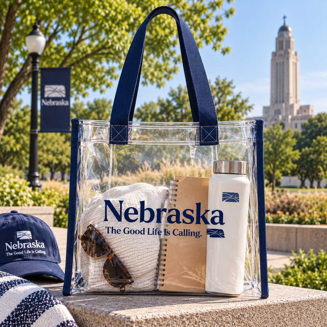 A clear tote bag with navy blue handles and trim, printed with the Nebraska state logo and slogan, sits on a stone bench outdoors. Inside the bag are a cream knit sweater, a brown notebook, and a white insulated bottle.