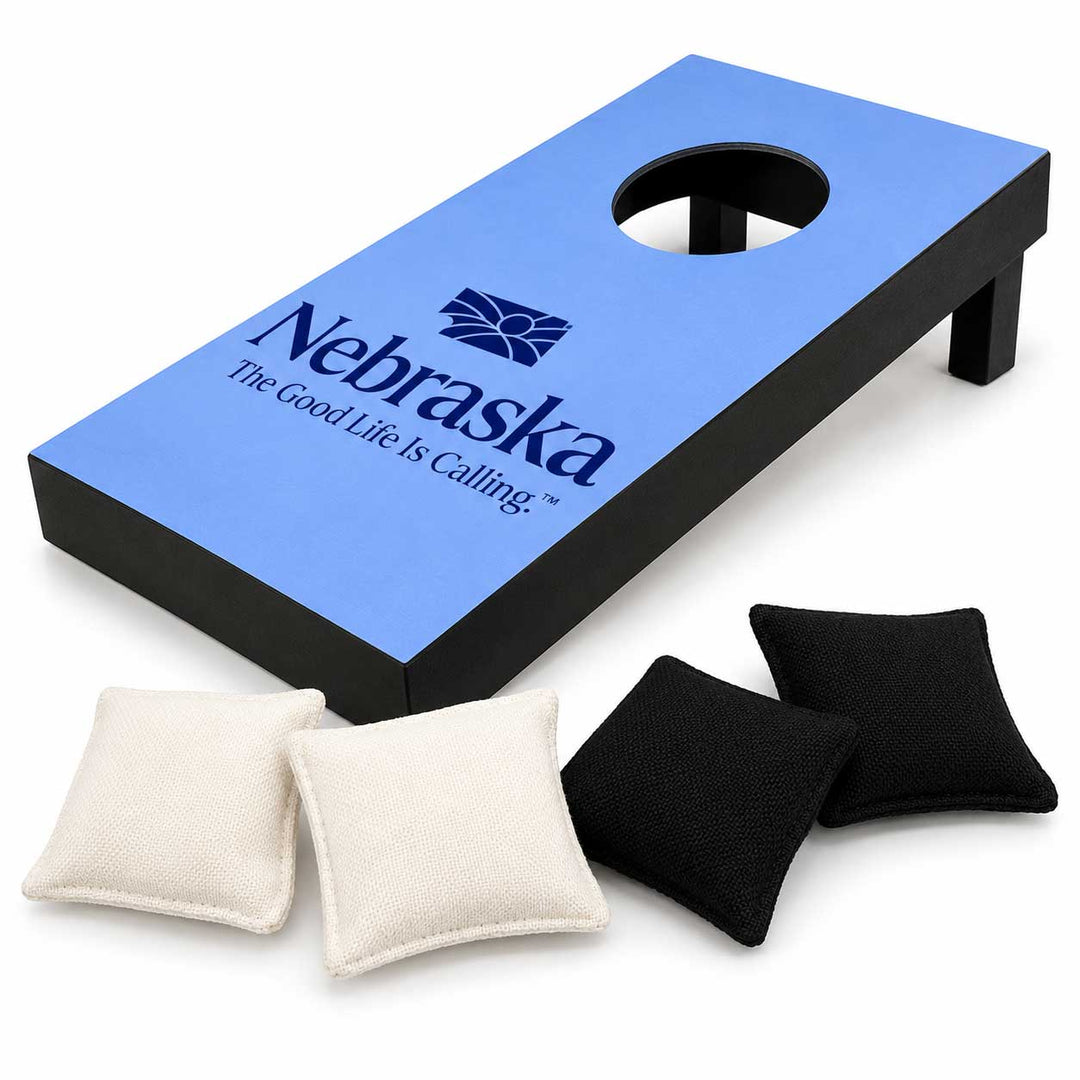 A cornhole board set with a blue and black board featuring the Nebraska logo, accompanied by two cream-colored bean bags and two black bean bags on a white background.