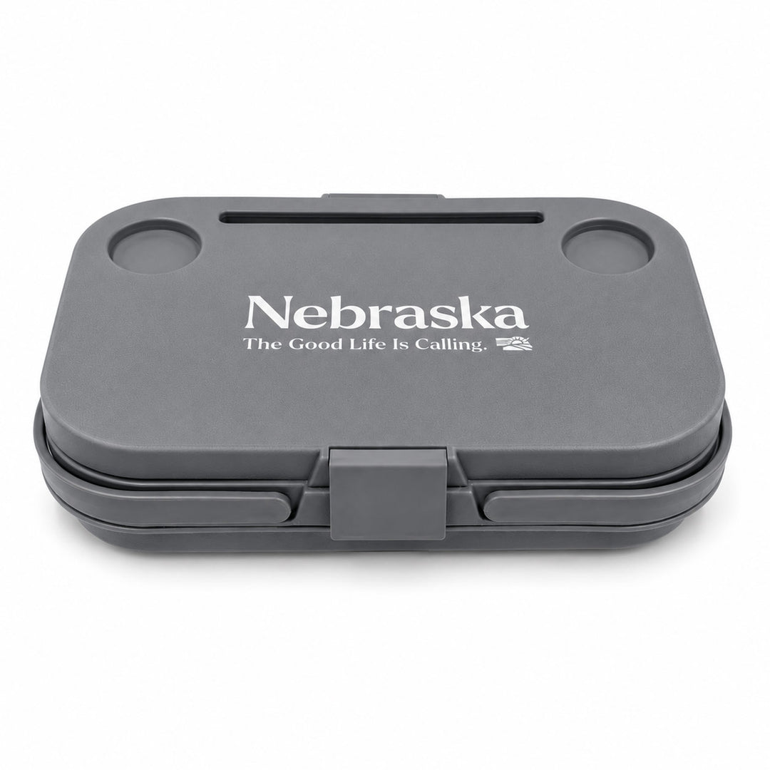 A dark gray hard-shell lunch box with a hinged lid, featuring the white printed text 'Nebraska' and 'The Good Life Is Calling.' on the lid, set against a plain white background.