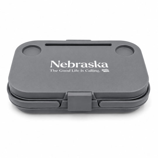 A dark gray hard-shell lunch box with a hinged lid, featuring the white printed text 'Nebraska' and 'The Good Life Is Calling.' on the lid, set against a plain white background.