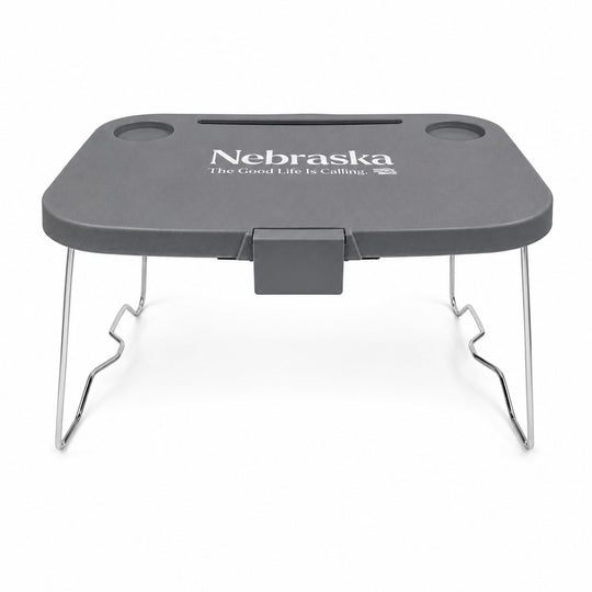 A dark gray portable lap desk with silver metal legs, featuring the white printed text 'Nebraska The Good Life Is Calling' on the top surface.