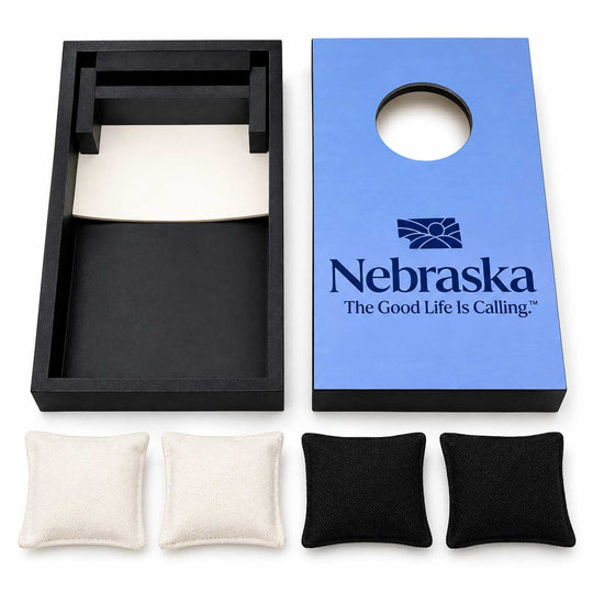 Nebraska cornhole set with a blue wooden board printed with the Nebraska logo, a black plastic hole cover, four fabric beanbags (two black and two off-white), and a black storage tray on a white background.