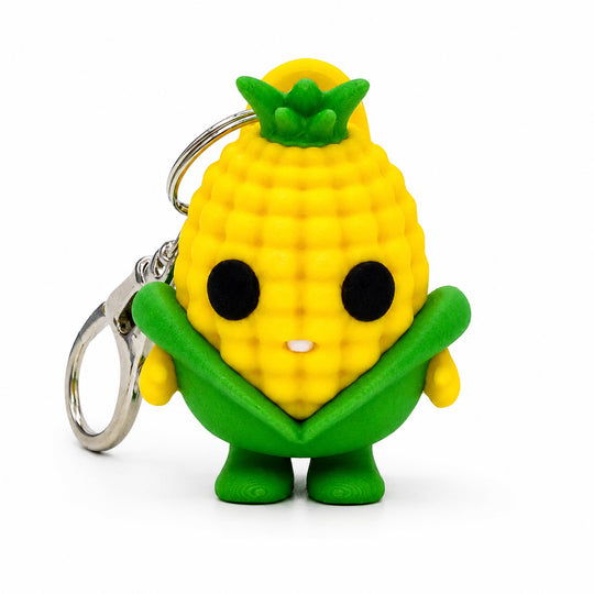 A cute, stylized corn keychain figure made of textured yellow plastic with green leafy accents, large black eyes, and a silver metal keyring, photographed on a clean white background.