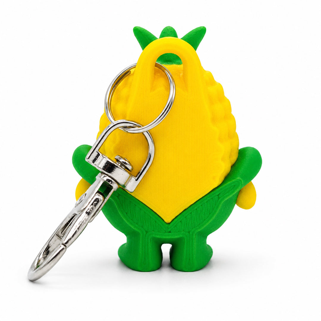 A novelty keychain shaped like a cartoon corn cob, featuring a bright yellow body, green leaves on top, green stem-like accents, and a silver metal split ring and clasp, shown on a plain white background.