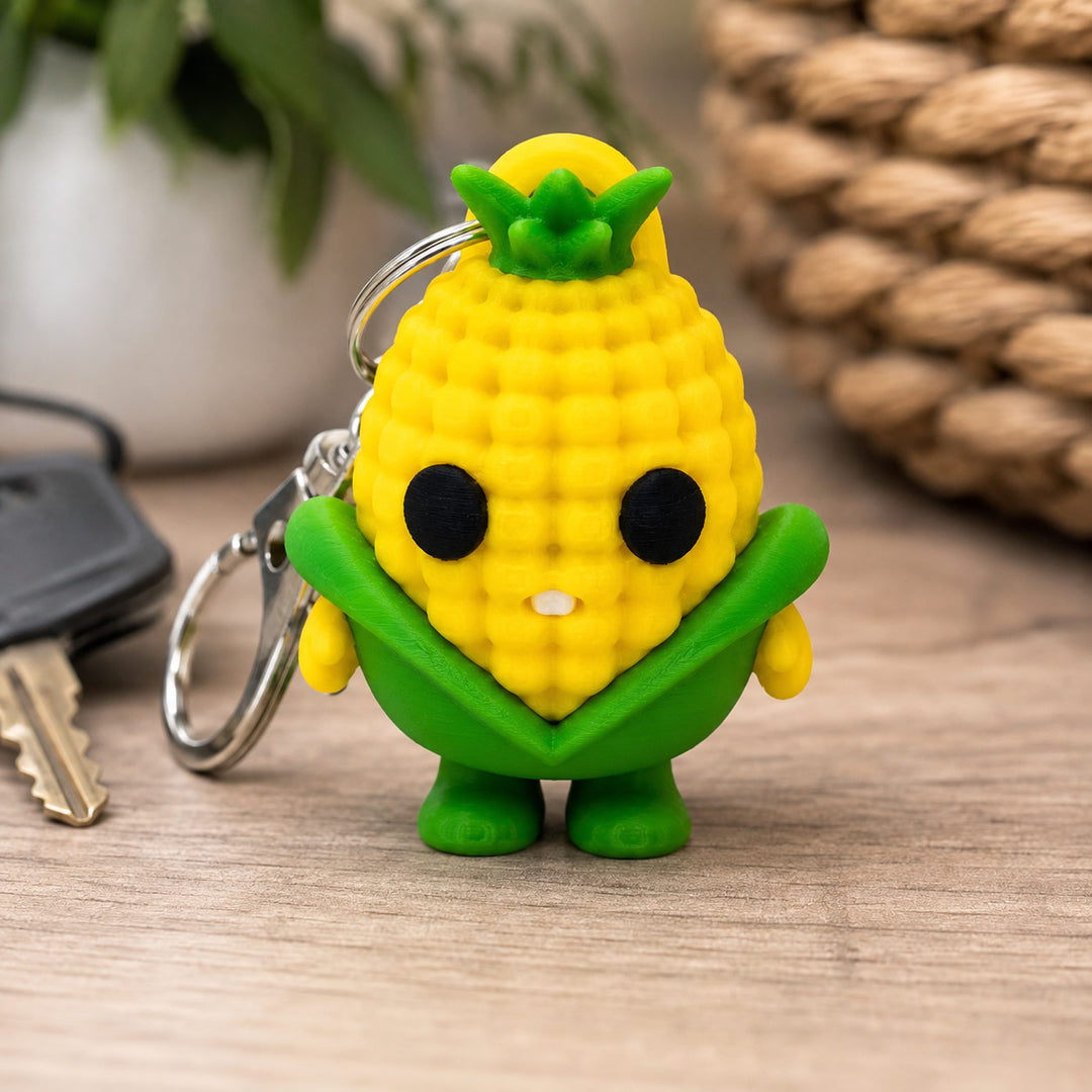 A small yellow and green corn-shaped keychain with a cute face, attached to a silver key ring, standing on a wooden surface with keys and a woven basket in the background.