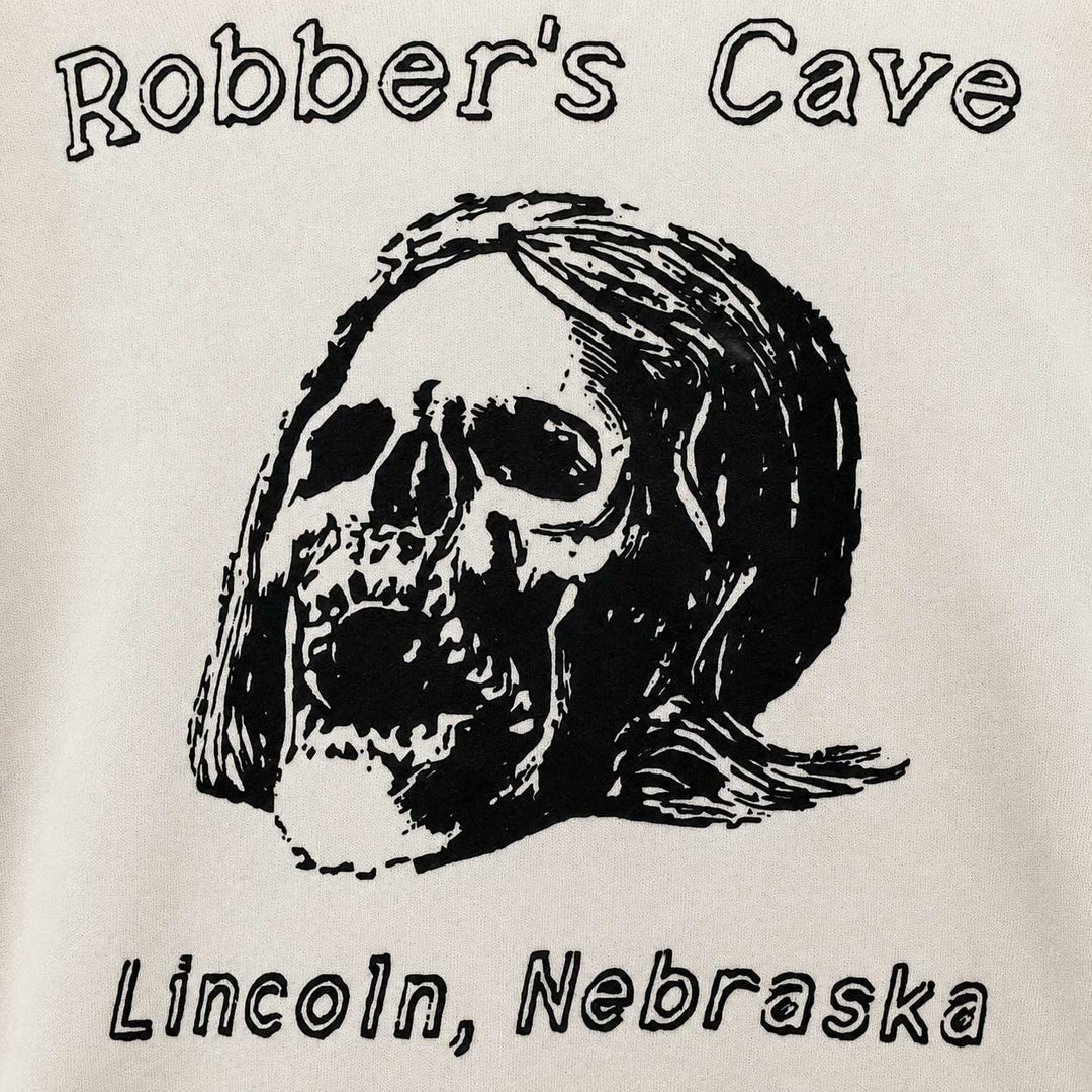 A close-up of a graphic print on a light-colored fabric, featuring a black-and-white illustrated skull with long hair, the words 'Robber's Cave' above, and 'Lincoln, Nebraska' below.