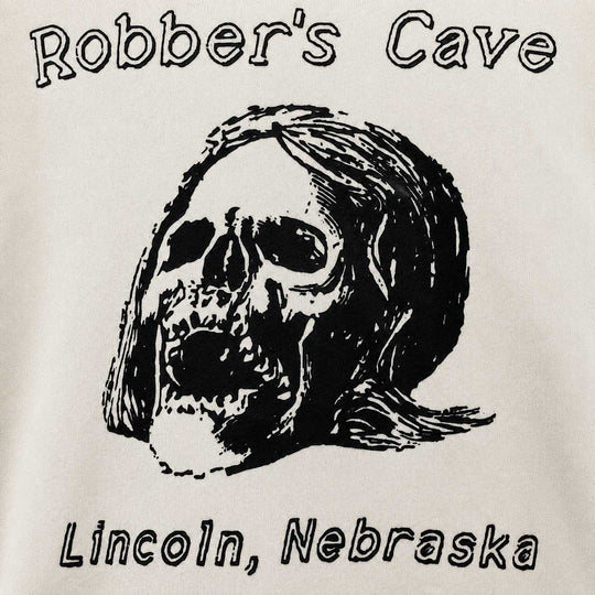 A close-up of a graphic print on a light-colored fabric, featuring a black-and-white illustrated skull with long hair, the words 'Robber's Cave' above, and 'Lincoln, Nebraska' below.