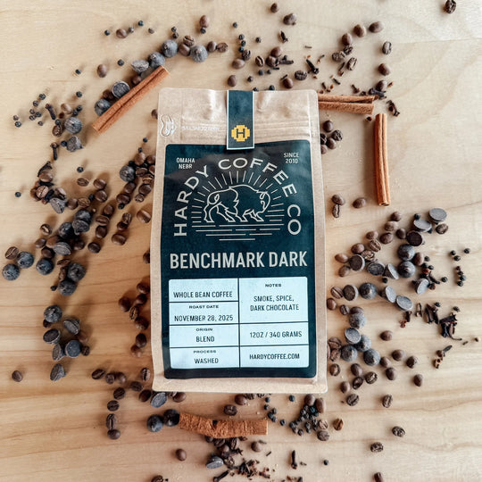 Hardy Coffee Co. Benchmark Dark coffee package on a wooden surface with coffee beans and cinnamon sticks.