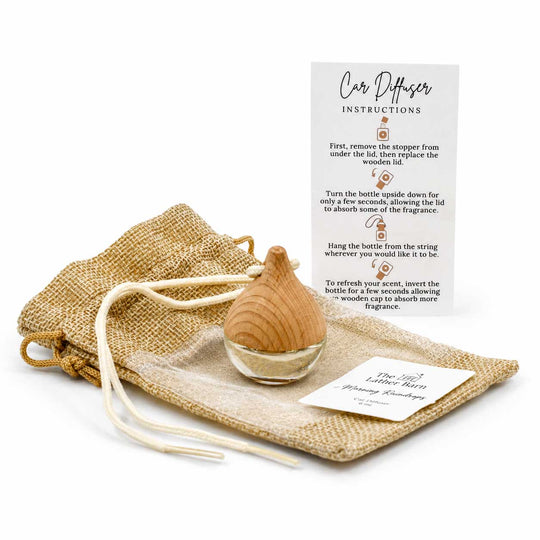 A wooden car diffuser bottle with a clear glass base, displayed with a burlap drawstring pouch, a printed instruction card, and a small product tag on a white background.