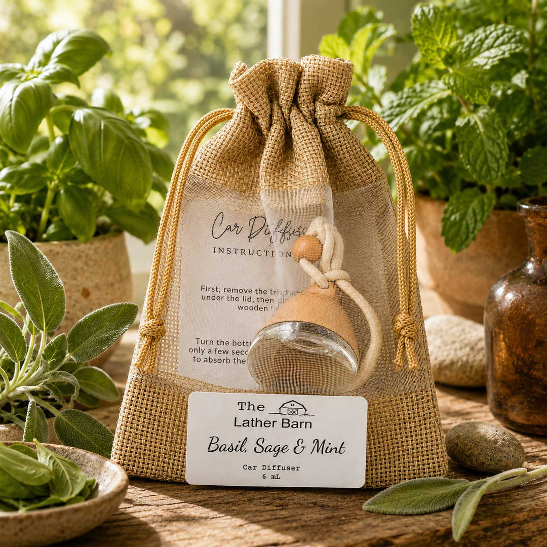 A car diffuser bottle in a clear mesh drawstring bag with burlap trim, labeled 'The Lather Barn Basil Sage & Mint Car Diffuser.' The bag sits on a wooden surface with basil leaves, herbs, and a rustic bottle in the background.