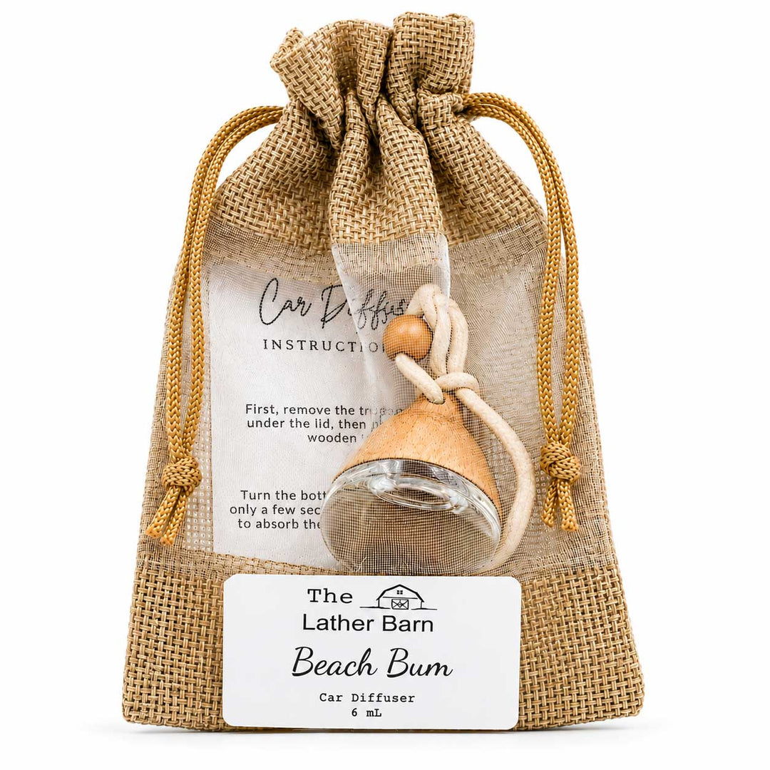 A small car diffuser bottle with a wooden bead and white cord, packaged inside a clear drawstring burlap pouch with a printed instruction card and a front label reading 'The Lather Barn Beach Bum Car Diffuser 6 ml'.