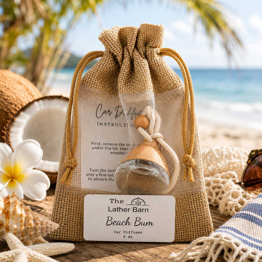 A car diffuser bottle in a clear glass bottle with a wooden diffuser cone, presented in a sheer beige burlap drawstring pouch with a printed instruction card and a white label reading 'The Lather Barn Beach Bum Car Diffuser 6 mL'. The scene is styled on a beach-themed surface with a coconut, white plumeria flower, starfish, and a blue-and-white striped towel, with a blurred beach background.