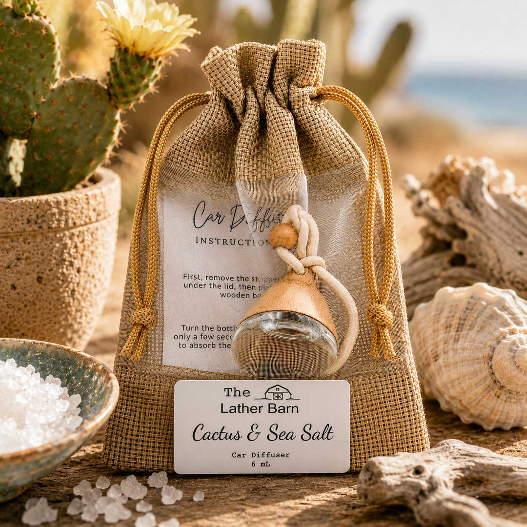A burlap drawstring pouch containing a small glass car diffuser bottle with a wooden diffuser top, displayed on a beach-themed surface with cactus, sea salt, and seashells. The pouch has a clear front label reading 'Car Diffuser Instructions' and a front tag for 'The Lather Barn Cactus & Sea Salt Car Diffuser'.