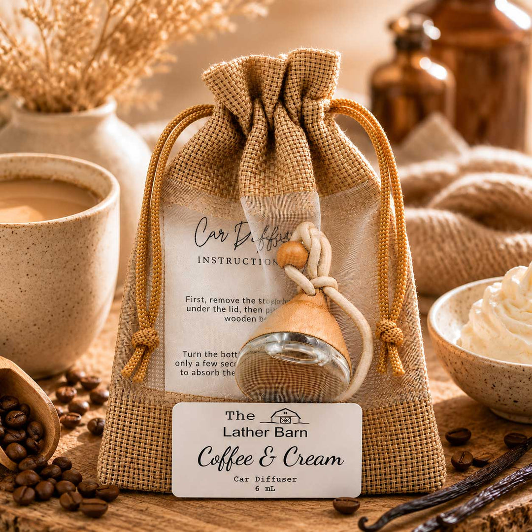 A small glass car diffuser bottle with a beige wooden collar, presented inside a burlap drawstring pouch with a printed instruction card and a front label reading 'The Lather Barn Coffee & Cream Car Diffuser.' The scene is styled on a wooden surface with coffee beans, a mug, whipped cream, and warm rustic props.