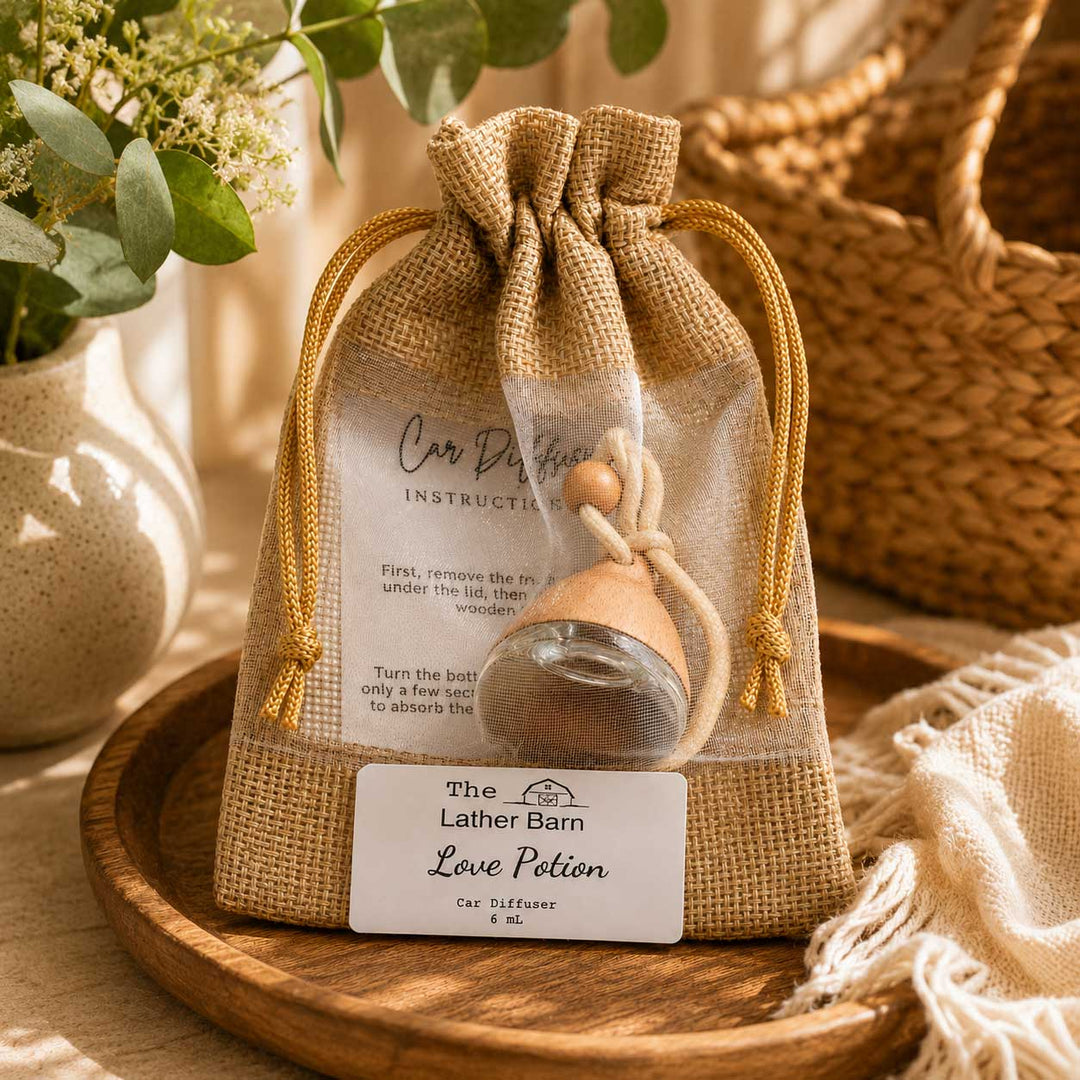 A clear drawstring pouch with a burlap-style front panel holds a small glass car perfume diffuser with a wooden stopper. A printed instruction card is visible inside, and a white label at the bottom reads 'The Lather Barn Love Potion Car Diffuser 6 ML.