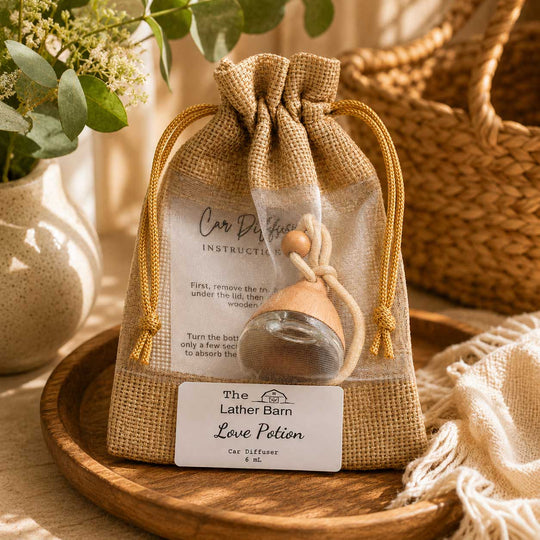 A clear drawstring pouch with a burlap-style front panel holds a small glass car perfume diffuser with a wooden stopper. A printed instruction card is visible inside, and a white label at the bottom reads 'The Lather Barn Love Potion Car Diffuser 6 ML.
