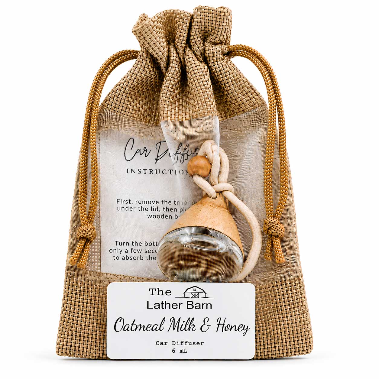 A small glass car diffuser bottle with a wooden top and cream cord, presented inside a clear drawstring burlap pouch with a printed insert and a front label reading 'The Lather Barn Calmant Milk & Honey Car Diffuser 6 mL'.