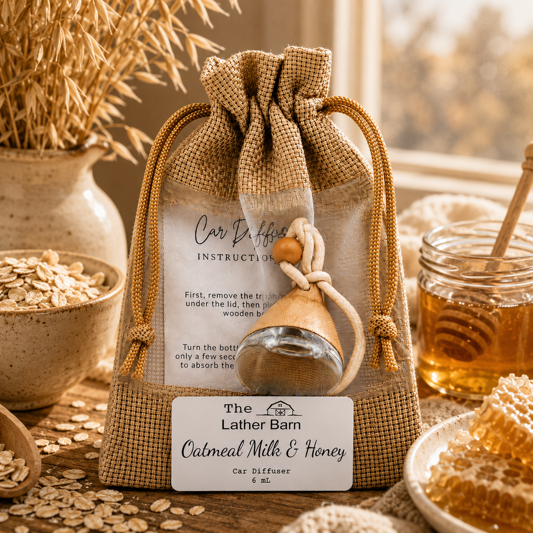 A burlap drawstring gift bag containing a small glass bottle of Carv Diffuser oil, labeled 'The Lather Barn Car Diffuser Oil' with an instruction card, styled on a table with oats, honey, and dried grass in a warm rustic setting.