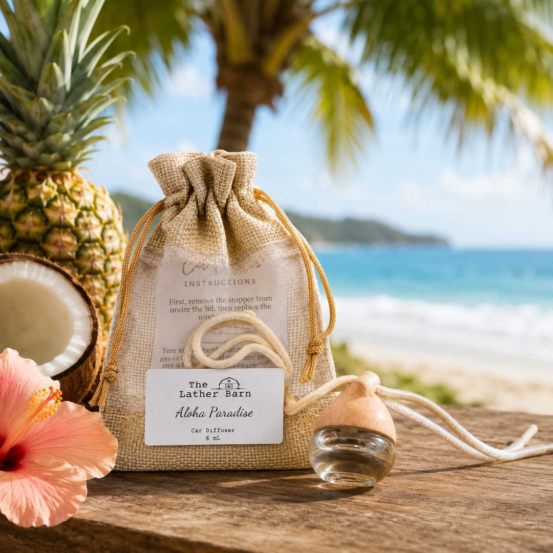A small burlap drawstring pouch with a white product label sits on a wooden surface, styled with a pineapple, a halved coconut, a pink hibiscus flower, and a small glass bottle with a cork stopper. The background is a blurred tropical beach scene with palm trees and ocean water.