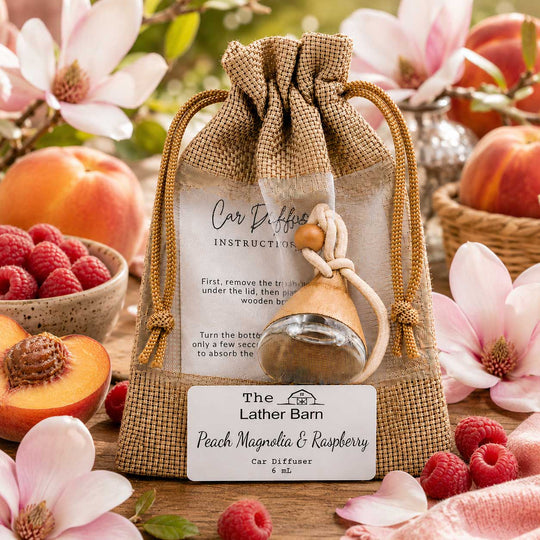 A burlap drawstring pouch containing a small glass car diffuser bottle with a wooden diffuser and a peach-shaped stopper, displayed on a wooden surface with peaches, raspberries, pink flowers, and an instruction card. The front label reads 'The Lather Barn Peach Magnolia & Raspberry Car Diffuser'.