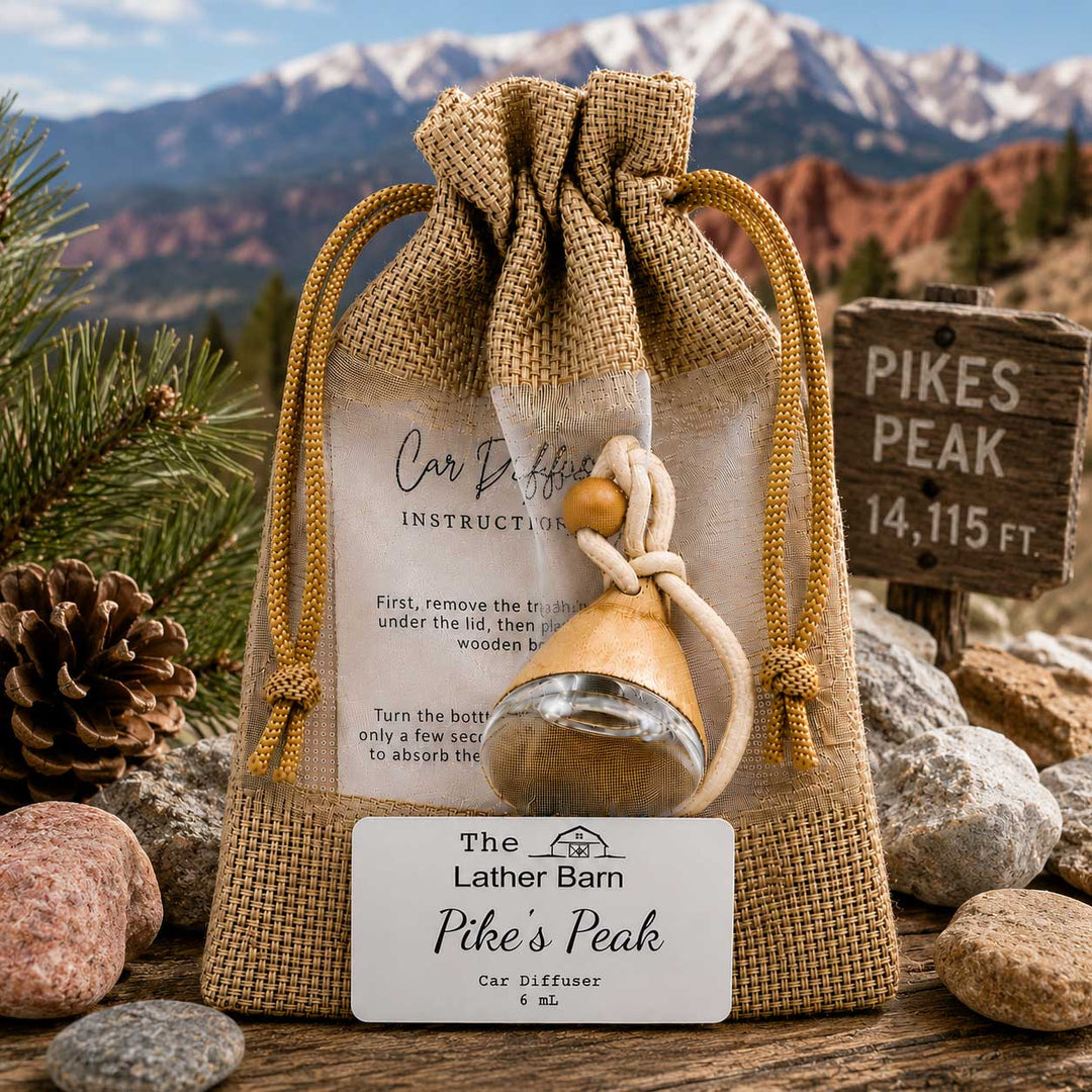 A burlap drawstring pouch containing a small wooden car diffuser bottle with a clear glass bottom, shown outdoors on a rustic wooden surface with rocks, pine branches, a pinecone, a sign reading Pikes Peak, and a mountain landscape in the background. A front label reads 'The Lather Barn Pike's Peak Car Diffuser'.