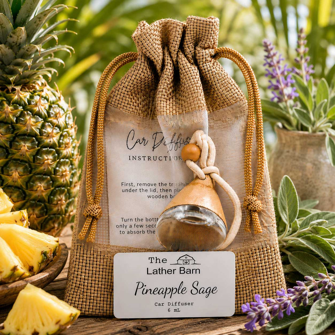 A burlap drawstring gift pouch containing a small glass car diffuser bottle with a wooden stopper, displayed with an instruction card and a front label reading 'The Lather Barn Pineapple Sage Car Diffuser 5 ml.' The scene is styled with pineapples, sliced pineapple, and greenery in an outdoor garden setting.