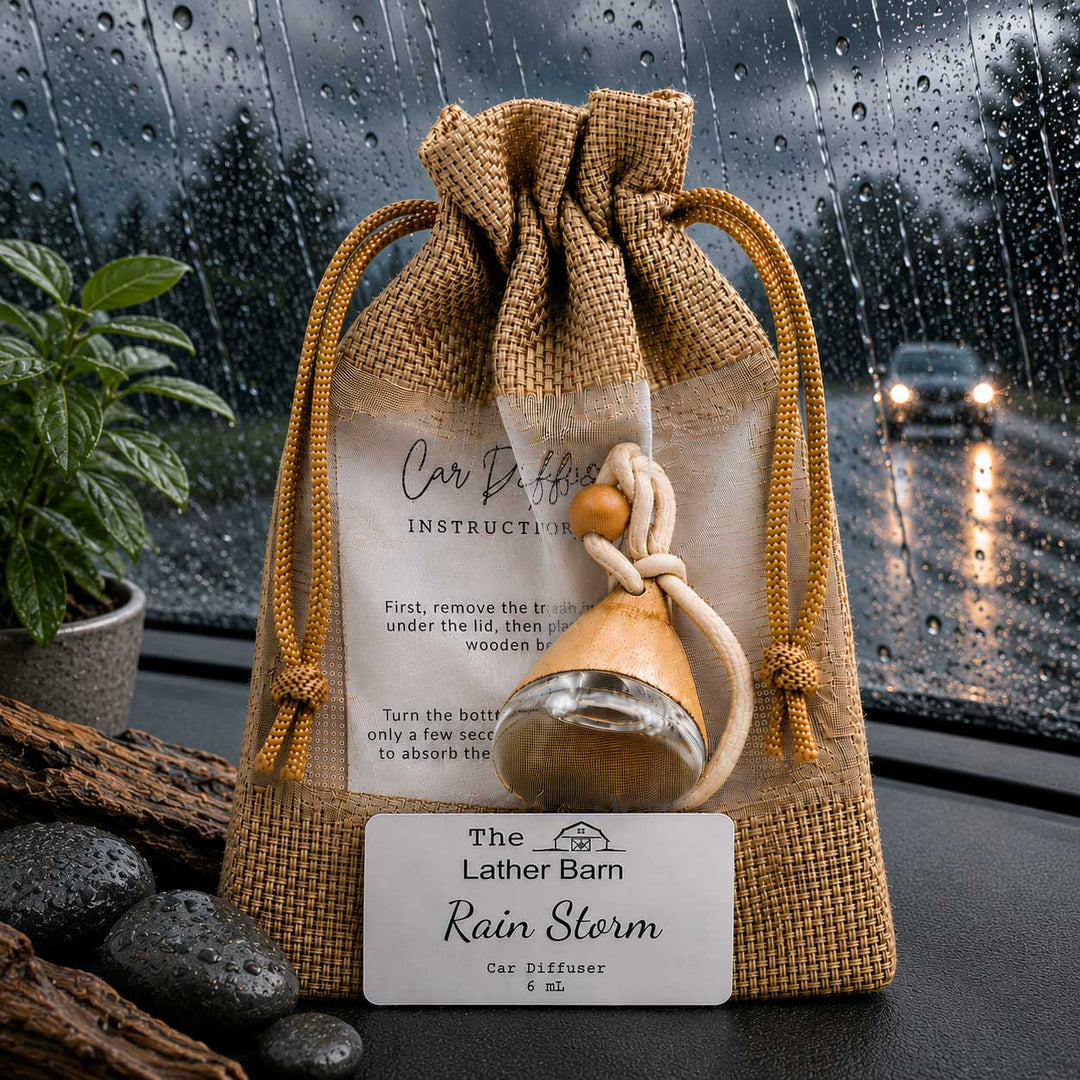 A burlap drawstring gift pouch with a clear front window displaying a small glass car diffuser bottle and printed instruction card. A white brand label reading 'The Lather Barn Rain Storm Car Diffuser' is placed in front.
