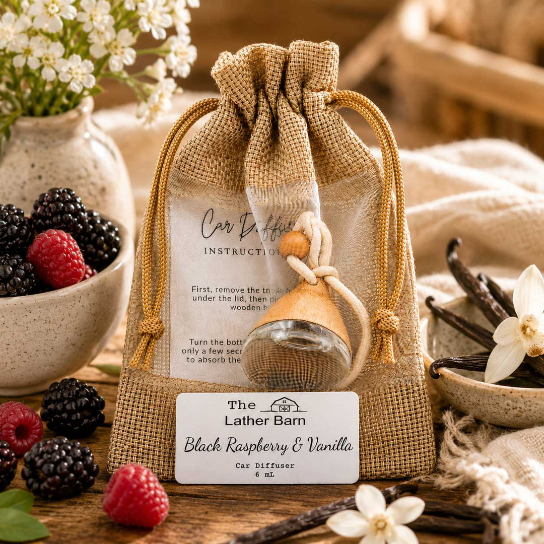 A burlap drawstring pouch containing a small glass car diffuser bottle with a wooden diffuser key, displayed on a rustic table with flowers, berries, vanilla beans, and a branded front label reading 'The Lather Barn Black Raspberry & Vanilla Car Diffuser 6 oz.'.