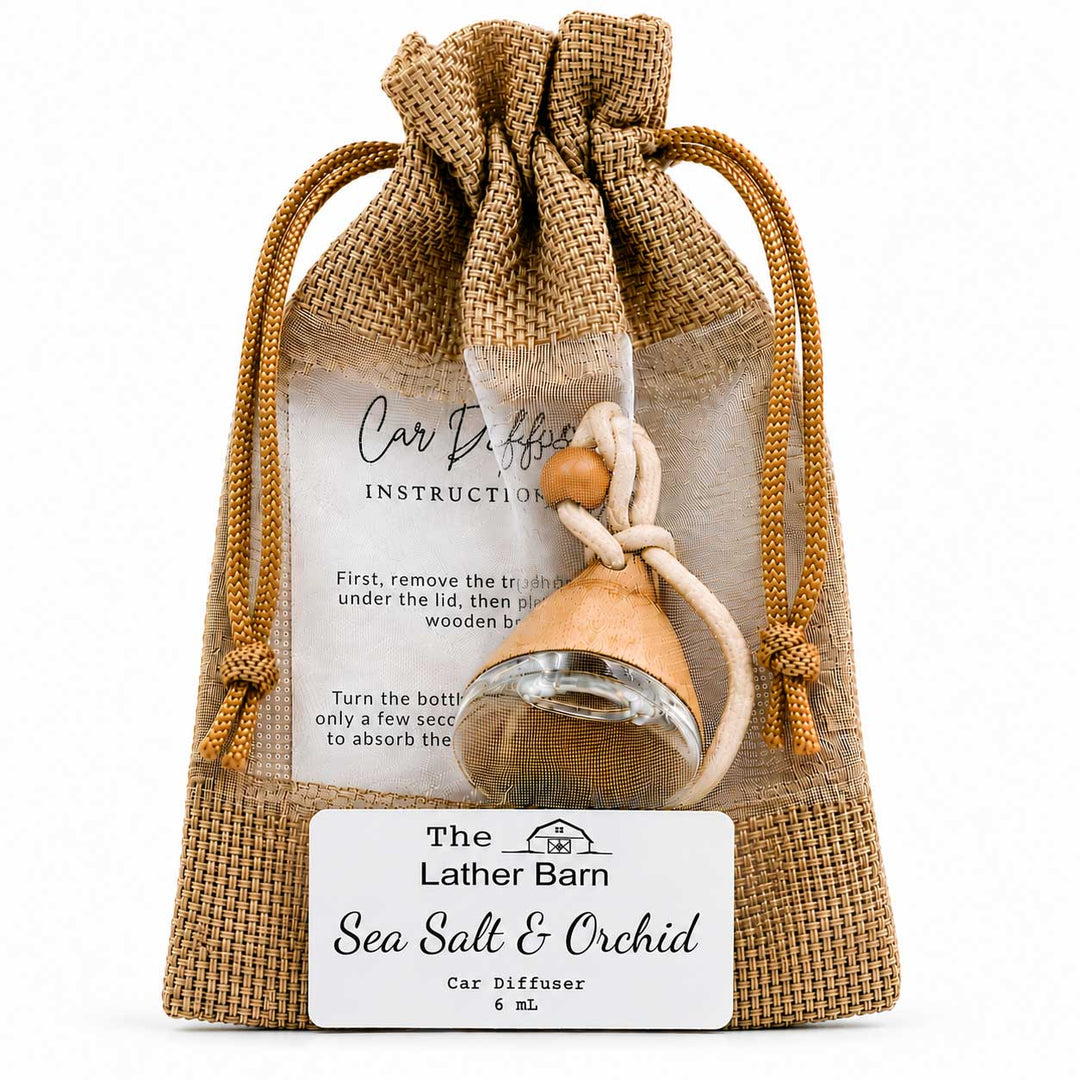 A car diffuser in a clear drawstring pouch with burlap trim, labeled 'The Lather Barn Sea Salt & Orchid Car Diffuser 6 mL.' A small wooden diffuser bottle with a rope tie and clear glass bowl is visible through the pouch, with an instruction card behind it on a white background.
