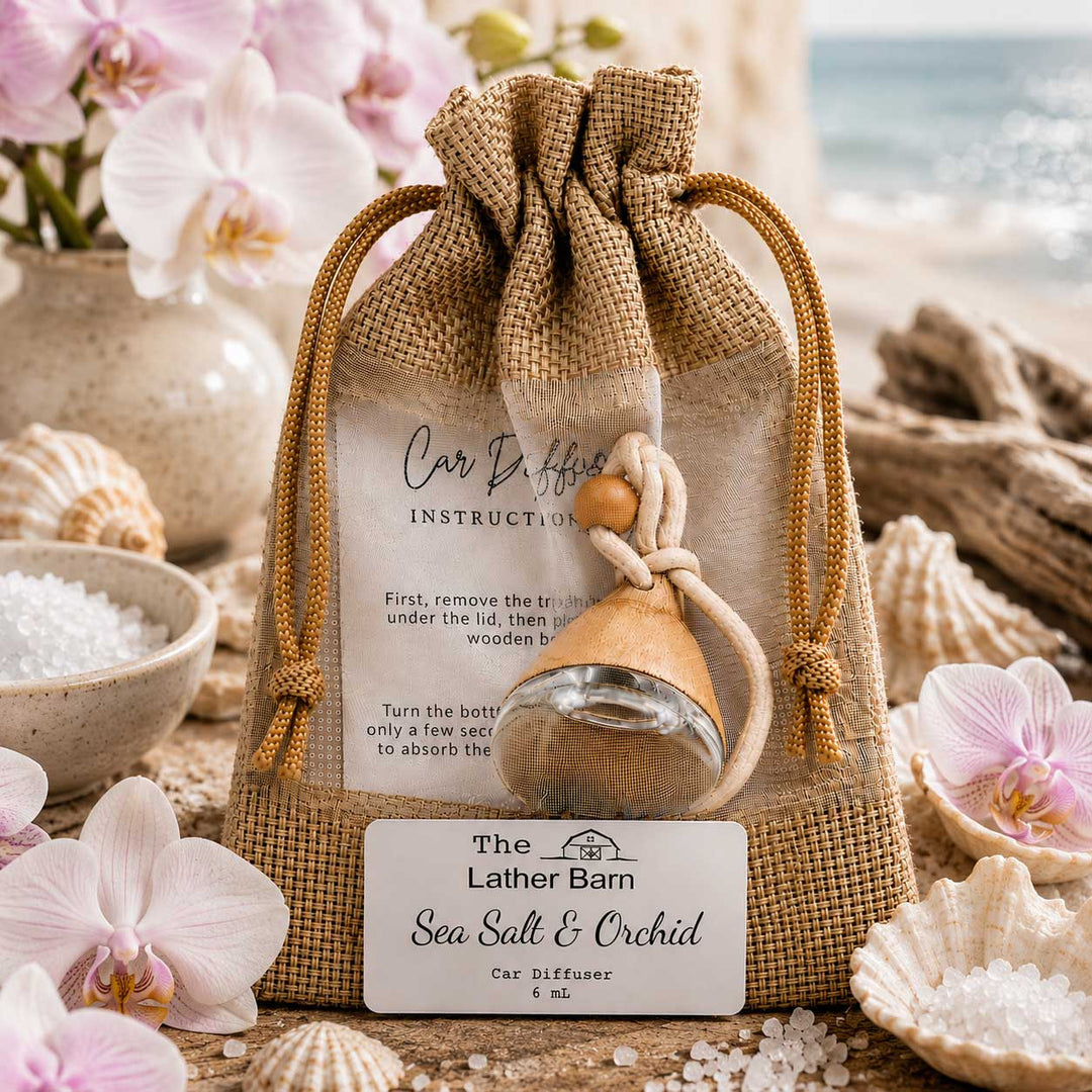 A burlap drawstring pouch containing a small glass car diffuser bottle with a wooden sprayer, displayed with a front label reading 'The Lather Barn Sea Salt & Orchid Car Diffuser.' The scene is styled with pink orchids, seashells, bowls of white salt, and a blurred beach background, making it a lifestyle product shot.