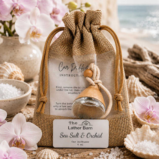A burlap drawstring pouch containing a small glass car diffuser bottle with a wooden sprayer, displayed with a front label reading 'The Lather Barn Sea Salt & Orchid Car Diffuser.' The scene is styled with pink orchids, seashells, bowls of white salt, and a blurred beach background, making it a lifestyle product shot.