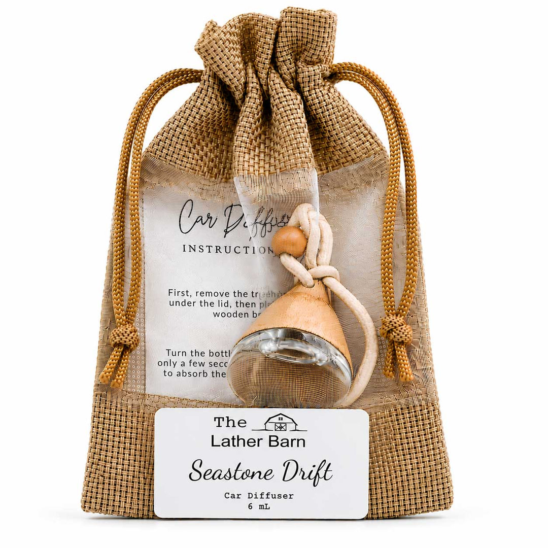 A car diffuser set in a burlap drawstring pouch with an instruction card and a white brand label. The diffuser features a small glass bottle with a beige seashell-shaped atomizer and a wooden turn key.