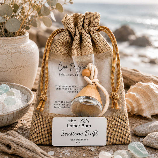 A burlap drawstring gift bag with a small car diffuser bottle tied inside, placed on a beach-style surface with shells, driftwood, and a blurred ocean background. A front label reads 'The Lather Barn Seastone Drift Car Diffuser.