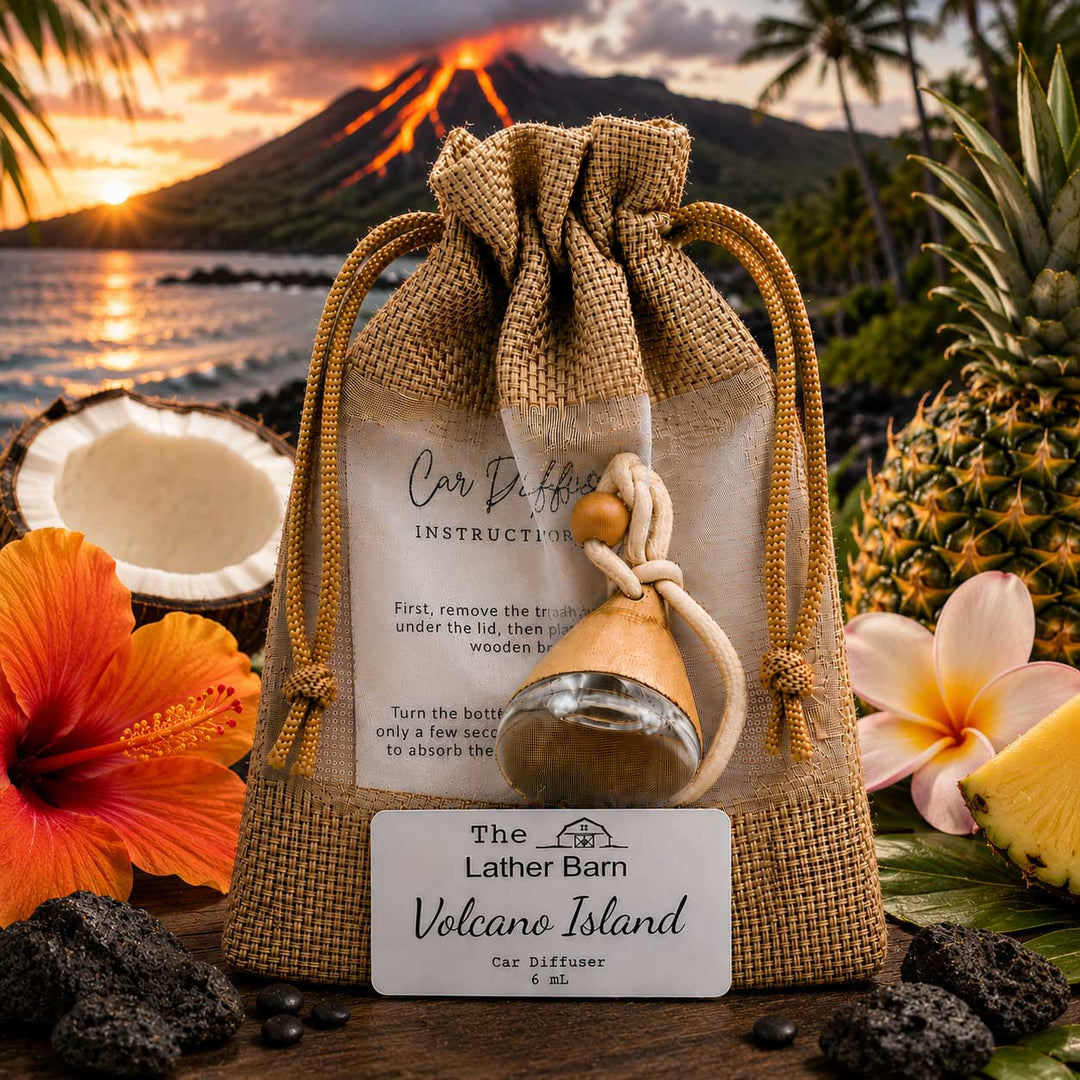 A car diffuser set displayed on a beach-themed surface with tropical flowers, coconuts, pineapples, rocks, and a volcano in the background. The product includes a burlap drawstring bag, an instruction card, a small glass bottle with a wooden stopper, and a branded card reading 'The Lather Barn Volcano Island Car Diffuser 6 lit'.