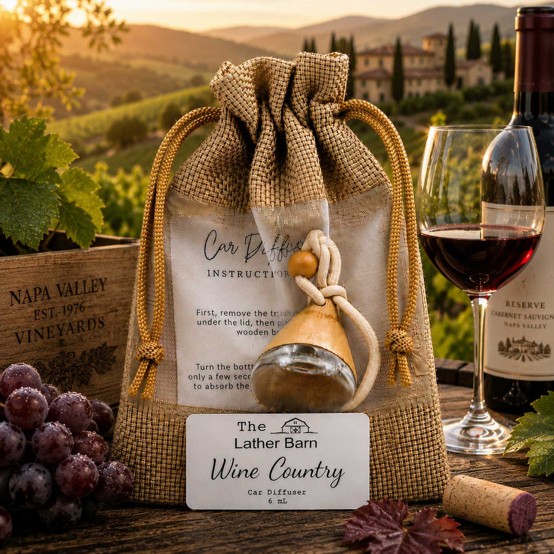 A burlap drawstring gift bag with a wine car distiller kit inside, displayed on a rustic wooden table with a wine glass of red wine, grapes, a cork, and a wine bottle in the background against a vineyard landscape at sunset.