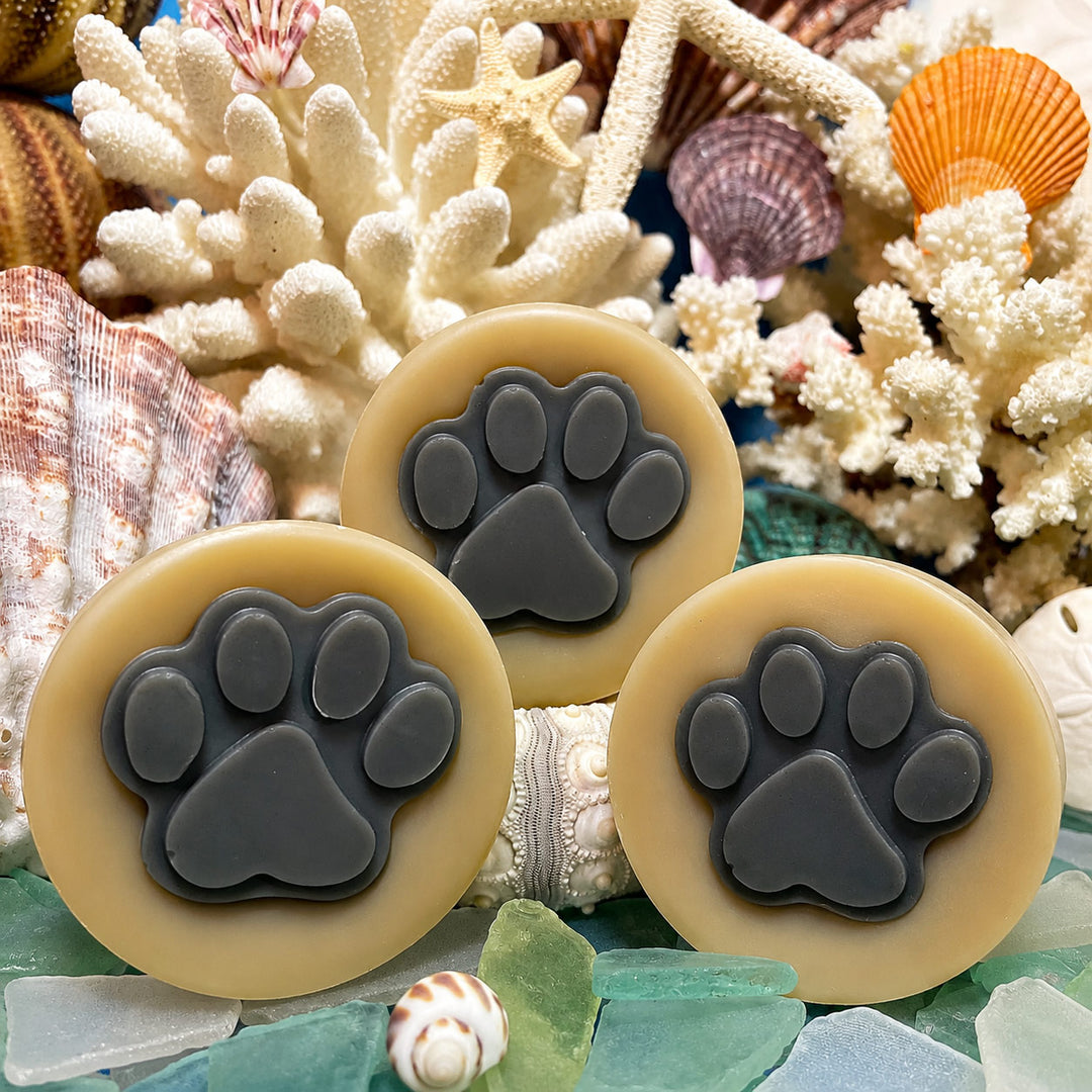 Three paw print-shaped soaps with beige and gray colors on a background of coral and shells.