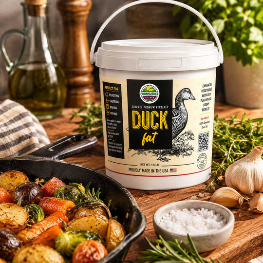 A white plastic tub of Cornhusker Kitchen Duck Fat seasoning mix sits on a rustic wooden table surrounded by cooking ingredients and prepared food. A cast-iron skillet with roasted vegetables, a small bowl of coarse salt, garlic, olive oil, and herbs frame the product, which features a label with a duck illustration and the Grasslands logo.