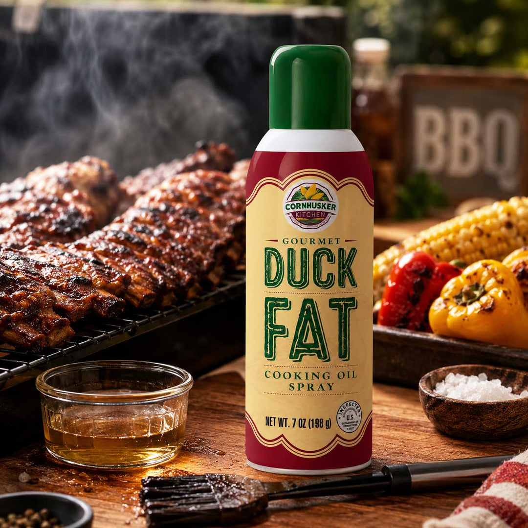 A can of Cornhusker Gourmet Duck Fat Cooking Oil Spray stands on a wooden table in a barbecue setting, with grilled meat, corn, peppers, salt, and a brush in the background.