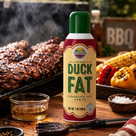 A can of Cornhusker Gourmet Duck Fat Cooking Oil Spray stands on a wooden table in a barbecue setting, with grilled meat, corn, peppers, salt, and a brush in the background.